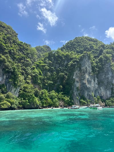 A wide-angle, eye-level shot captures a tropical scene of verdant, jungle-covered cliffs rising steeply from a turquoise sea. The cliffs are rugged and grey, with streaks of brown and white running down their faces. Lush green vegetation clings to the steep slopes and rocky outcrops. The water in the foreground is crystal clear, revealing the sandy bottom in shallower areas. Several boats are anchored near a small, sandy beach nestled at the base of the cliffs. People can be seen on the beach and on some of the boats. The sky above is a bright blue, dotted with fluffy white clouds. The overall impression is one of a serene and beautiful natural landscape. This scene is located in the city of Haad Hin Khom, Thailand.