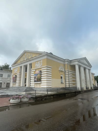 The image shows the Zavolzhsky City Drama Theater in Zavolzhye, Russia, on an overcast day. The theater is a grand, neoclassical building with a yellow facade, white columns, and decorative friezes. A wide staircase leads up to the entrance. In front of the theater, there is a black metal fence bordering a wet street with puddles reflecting the building and the sky. Two white planters filled with pink and purple flowers are situated on the sidewalk to the left of the stairs. Posters are displayed on the walls of the theater, indicating current performances or attractions. The overall scene suggests a somewhat damp and quiet atmosphere, characteristic of a cloudy day in a city square.