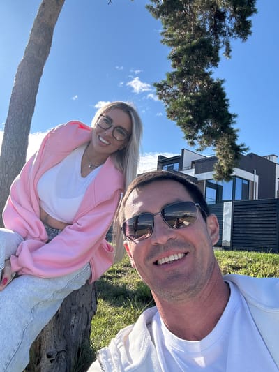 This is a selfie of a man and a woman outdoors, with a modern house and trees in the background. The man is in the foreground, smiling and wearing sunglasses and a white jacket. The woman is behind him, wearing a pink hoodie and glasses, also smiling. The scene appears to be a sunny day. Based on the architectural style and landscaping, this location could be in a residential area, possibly with a view of the sea or a body of water, which is typical of many areas in Primorsky Krai, Russia.