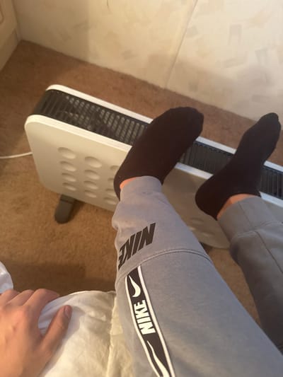 In an indoor setting within Fowlis Easter, United Kingdom, a close-up perspective captures the lower body of an individual. The person is wearing grey sweatpants featuring black "NIKE" branding on the left thigh and a black and white vertical stripe with the "NIKE" logo and text on the outer left leg. Both feet are clad in black ankle socks and are positioned elevated above the top grille of a white electric fan heater. The heater, with a black top grille and front circular indentations, rests on a light brown carpeted floor. A white power cord extends from the heater's left side. In the lower-left foreground, the person's left hand rests on a crumpled white surface, likely bedding or a cushion. A light-colored wall with subtle rectangular patterning is visible in the background, including a corner.