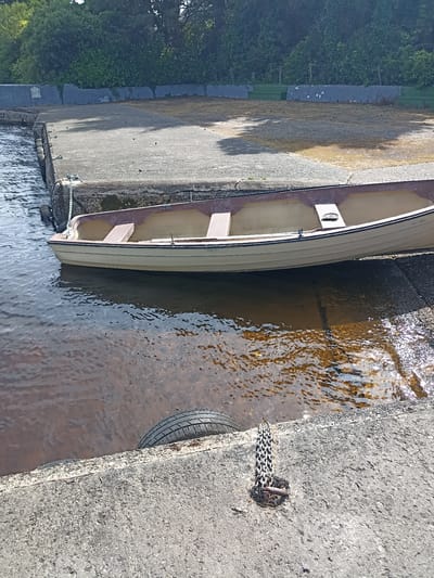 The image shows a tan and brown rowboat partially submerged in dark, murky water next to a concrete dock. The dock is weathered and cracked, with a tire visible along its edge. A metal chain is attached to the dock and dangles into the water. In the background, lush green trees and foliage are visible, with a clear blue sky above. The overall scene suggests a quiet, natural setting, likely a harbor or inlet.