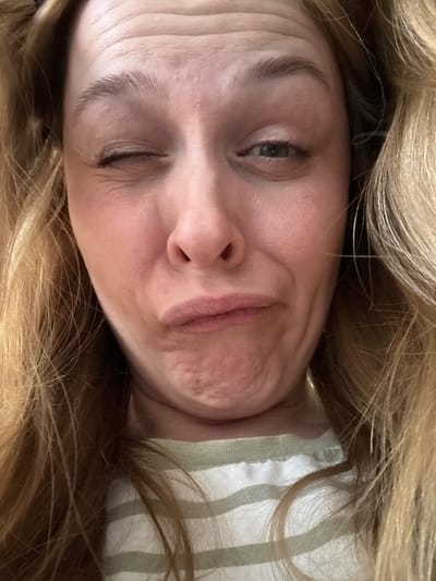 The media file is a close-up, vertically oriented photograph of a woman's face, framed by her long, blonde hair. The woman is looking directly at the camera with a slightly puckered expression, her lips pursed. Her right eye is partially closed, creating a wink-like effect, while her left eye is open and appears to be looking forward. Her skin has visible texture, with subtle wrinkles around her eye and forehead, suggesting a neutral or possibly playful emotion.
She is wearing a striped garment, possibly a shirt or blouse, with vertical stripes in light green and white. The fabric appears to be made of cotton or a similar material. The background is dominated by her hair, which is light blonde with some lighter and darker strands, suggesting natural highlights or variations in color.
The lighting is soft and diffused, indicating an indoor setting or an overcast day. There are no discernible location cues to pinpoint a specific place within Радваніцкі сельскі Савет, Belarus, other than the general setting of a person in casual attire, likely at home. The image is a still photograph and does not depict any ongoing activity or event beyond the posed expression of the subject. There is no visible text in the image.