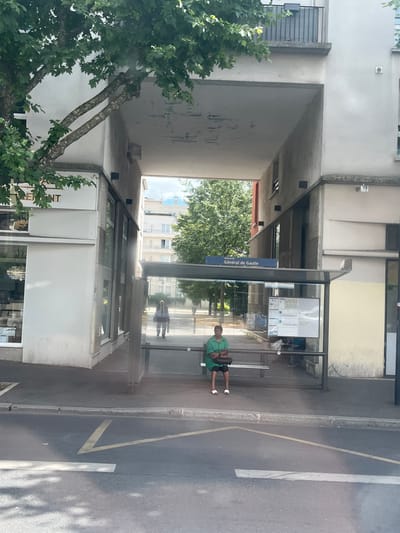 The image shows a street scene in Alfortville, France, specifically at a bus stop labeled "Général de Gaulle." In the foreground, there is a paved road with yellow markings. Behind the road is a sidewalk and a bus stop shelter. A person, identified as an adult, is seated on the bench inside the bus stop shelter, wearing a green shirt and dark pants, and white sandals. They are holding a dark bag on their lap. Reflected in the glass of the bus stop shelter, another person is visible walking away. The bus stop is situated in front of a multi-story building with balconies and windows. Overhanging the scene from the left is a large tree with green foliage. The background shows more buildings and trees under a bright sky.