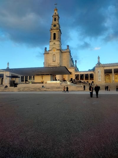 The image depicts the Sanctuary of Fátima in Fátima, Portugal, specifically the Basilica of Our Lady of the Rosary and its surrounding plaza, the Esplanada.
In the background, the Basilica of Our Lady of the Rosary stands centrally. It is a light-colored, ornate stone building topped by a tall bell tower featuring a clock face, arched openings, and a crown and cross at its apex. Below the bell tower, a niche on the basilica's facade contains a white statue. Sculptural elements are visible along the roofline.
In the mid-ground, a large, dark, flat canopy structure extends forward from the basilica's base, covering an elevated stage or altar area. A depiction of a crucifix is visible on the front of this covered stage. Wide, light-colored stone steps lead up from the Esplanada to the basilica and the covered stage. To the right of the basilica, a colonnaded wing extends, also constructed of light-colored stone, with banners visible on its facade.
The foreground is dominated by a vast, dark gray paved square, the Esplanada, which appears largely unadorned.
Several individuals are present within the scene. In the central foreground, two individuals are walking side-by-side, moving away from the viewer. One wears dark clothing, the other a light brown coat. Further to the left on the Esplanada, two additional individuals are walking. Numerous smaller figures are observed ascending and descending the wide stone steps leading to the basilica, and some are visible under the colonnaded structure. One individual is seated on a step near the center.
The sky above features a gradient of pale blue towards the horizon, transitioning into darker, dense cloud formations overhead and towards the right, indicating conditions other than clear skies. The lighting suggests either late afternoon or early morning.