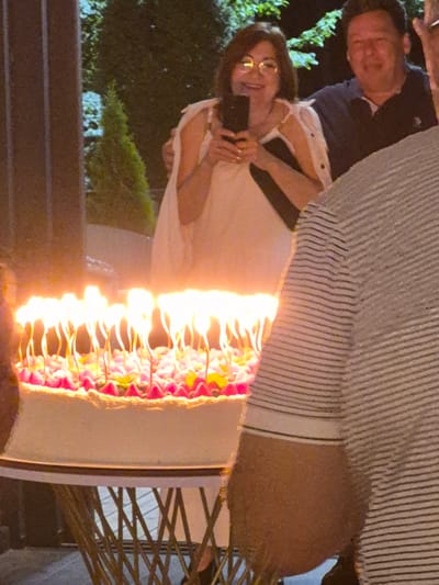 The image shows a birthday cake with lit candles on a table. A woman in a white dress is holding a phone and smiling. A man in a dark shirt is standing behind her. The scene appears to be outdoors at night, with trees and foliage in the background. The exact location in Zelyonovka, Russia, is not specified in the image.
