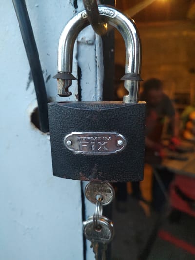 A close-up, eye-level shot captures a weathered, black, rectangular padlock with a silver metal plate on its front. The plate is embossed with the words "PREMIUM FIX" in an arch above the word "FIX" in larger letters. The padlock's silver shackle is partially open, revealing its locking mechanism. Two keys hang from a metal ring attached to the bottom of the padlock. The padlock is secured to a light blue metal surface, likely a door or gate, which shows signs of rust around a circular hole near the shackle. To the left of the padlock, a dark gray, cylindrical rod runs vertically. The background is blurred and appears to be an indoor or semi-outdoor setting with warm lighting and indistinct figures of people, suggesting a workshop or storage area. The overall impression is one of an outdoor, possibly neglected, urban environment in Kochmar, Bulgaria.