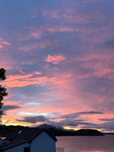 The image depicts a serene twilight scene in Sconser, United Kingdom, overlooking a body of water. The sky is a canvas of vibrant colors, with hues of pink, orange, and purple dominating the clouds, suggesting the last moments of sunset or the approaching dawn. In the foreground, the rooftops of two houses are visible, one with dark grey tiles and a small skylight, the other with lighter colored walls. The houses appear to be situated close to the water's edge. Beyond the houses, a dark silhouette of rolling hills and mountains stretches across the horizon. The water reflects the soft colors of the sky, creating a peaceful and atmospheric ambiance. The overall impression is one of tranquility and natural beauty in a coastal or lakeside setting.