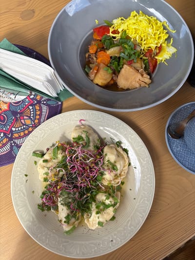 This is an overhead shot of two plates of food on a wooden table. The plate in the foreground is a light cream color with an embossed floral pattern around the rim. It contains several pierogi topped with chopped parsley and a pile of pink and green sprouts. The second plate, a larger gray bowl, is in the background and contains a stir-fry with what appears to be chicken, potatoes, carrots, red peppers, and green onions, topped with shredded yellow cabbage. To the left of the foreground plate, there are folded white napkins on top of a green napkin and a patterned fabric coaster. To the right of the gray bowl, a blue saucer holds a blue cup with a wooden spoon inside. The setting appears to be a restaurant or cafe in Górka, Poland.