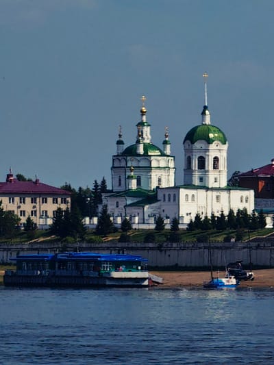 The image depicts a waterfront scene in the city of Yeniseysk, Russia. In the background, a large, white church with green domes and golden crosses stands prominently against a clear blue sky. Adjacent to the church is a building with a reddish-purple roof and multiple windows. In the foreground, a blue and white boat is anchored on a sandy shore, with a dark SUV parked nearby. Further to the left, a long, blue, multi-story boat or ferry is docked along the waterfront. The water in the foreground is a deep blue, reflecting the sky above. Trees are scattered throughout the landscape, dotting the hills behind the church and along the shoreline.