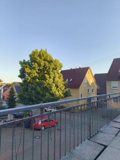 A high-angle, full outdoor shot captures a residential street in Hardthausen am Kocher, Germany, on a clear day. The sun illuminates the scene from the right, casting long shadows. In the foreground, a metal railing adorned with a string of decorative lights stretches across the bottom of the frame. Beyond the railing, a paved courtyard area is visible, with a red station wagon parked in the center. A dark gray SUV is parked to the left of the station wagon. A large, leafy green tree dominates the mid-ground, its branches reaching towards the bright blue sky. Several residential buildings with red-tiled roofs are visible behind the tree. The houses feature yellow and light beige facades, with windows and dormers. In the distance, power lines are visible against the sky. The overall atmosphere is peaceful and depicts a typical suburban environment.