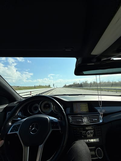 The image depicts the interior of a Mercedes-Benz car, likely while in motion on a highway near the city of Svoboda, Bulgaria. The view is from the driver's perspective, with the steering wheel prominently featuring the Mercedes-Benz logo. The dashboard is visible, showing a navigation screen with text in Bulgarian and some buttons. The speedometer indicates a speed exceeding 88 kilometers per hour. Beyond the windshield, a straight highway stretches into the distance under a blue sky with scattered clouds. The landscape beside the road appears to be a combination of grassy fields and trees. The image orientation is rotated, likely from being taken with a mobile device.