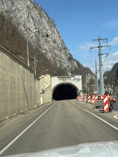 The scene depicts a clear daytime view of a two-lane asphalt road approaching the entrance of a tunnel within the city of Monastyr', Russia. The tunnel entrance features an arched dark opening framed by a light-colored, decorative stone facade. Above the arch, a sign displays Cyrillic text, reading "РОМАНТИК" above what appears to be "ТУР". To the immediate left of the road, a tall, grey concrete retaining wall with vertical panels runs alongside the base of a steep, rocky mountainside. Multiple streetlights on grey poles are mounted along the top edge of this wall and near the tunnel.
The mountainside, composed of grey rock with sparse, bare deciduous vegetation, rises sharply on both sides of the road and tunnel. On the right side of the road, a construction or maintenance area is delineated by several red and white striped traffic barricades. Behind these barricades, stacks of paving stones or similar materials are visible on pallets. Two individuals are present in this work zone: one person, wearing an orange high-visibility jacket, is partially obscured while bending over near the stacked materials; another person is less distinctly visible further back. A tall metal lattice electricity pylon, carrying multiple power lines, stands on the right side of the scene, with another smaller pylon partially visible behind it. The photograph is taken from inside a vehicle, with a portion of the dashboard or hood visible at the bottom of the frame. The sky above is clear and blue.