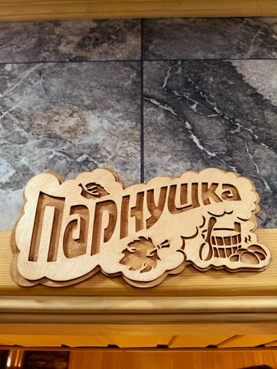 The image shows a wooden carved sign with Cyrillic lettering reading "Парнушка". The sign has a decorative cloud-like border and features images of leaves and what appears to be a bucket or barrel, suggesting a sauna or bathhouse theme. The sign is positioned horizontally on a wooden surface, with a marbled tile wall in the background. There are no people visible in the image. The location is identified as Kok Jar, Kyrgyzstan.