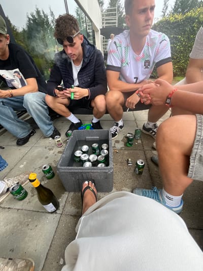 An outdoor scene captured from a low vantage point depicts at least five individuals on a paved patio or terrace, adjacent to a modern white building with large reflective windows. The location is an unspecified residential or private outdoor area within Skanderborg, Denmark.
In the immediate foreground, the lower legs and feet of an individual wearing white trousers and black flip-flops with green accents are visible; toes are painted with white nail polish.
To the left, a person wearing a dark baseball cap, black t-shirt with a graphic print, and light blue jeans is seated. A multi-colored bracelet is on their left wrist. Central in the frame, a young male with curly brown hair and round sunglasses, dressed in a dark quilted jacket and shorts, squats. He holds a black smartphone in his left hand and a neon green device (consistent with a vape pen) in his right hand, exhaling a plume of vapor from his mouth. To his right, another young male, wearing a white Portugal national football team jersey with a "Puma" logo and the number "7", also squats. He holds a lit cigarette in his right hand and observes something off-camera. A black wristband is on his left wrist. The lower body of a fourth individual, wearing light gray shorts and light blue athletic shoes, is visible on the right, with hands clasped and multiple red and black wristbands.
Centrally placed on the ground is a grey foam cooler containing water and approximately twelve to fifteen silver-colored beverage cans, partially submerged. Scattered on the light grey paved surface around the cooler are several green Carlsberg and Royal Export beer cans (some empty or crushed), a green glass bottle with a yellow foil top (consistent with sparkling wine), multiple cigarette butts, and a small black lighter. A blue and green plastic toy object rests near the cooler.
The background features the modern building with reflections of trees and sky in its large windows. Beyond the building, green foliage and a distant black patio umbrella are visible, along with a white structural element resembling scaffolding. The scene is illuminated by natural daylight.