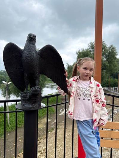 A young girl with blonde hair in pigtails stands next to a black eagle statue on a cloudy day. She is wearing a pink t-shirt with the words "All We Need Is Love" and a white jacket adorned with red hearts and other symbols. She is also wearing light blue pants. The eagle statue is large and dark, with its wings spread wide, and it is perched on a rock. Behind the girl and the statue, a river can be seen with trees and vegetation on the opposite bank. The scene appears to be an outdoor viewpoint, possibly a park or a scenic overlook, in Krasnoufimsk, Russia. The girl is looking at the camera with a neutral expression.