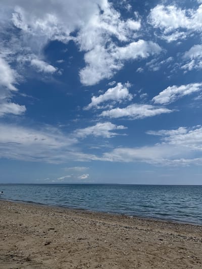 The image presents a landscape view of a beach in Νέα Φλογητά, Greece. The scene is composed of a sandy beach on the right, the Aegean Sea in the middle, and a sky filled with white clouds on the left. The sky transitions from a deep blue on the left to a lighter blue near the horizon over the sea. The sand on the beach is a light brown color, and some sparse vegetation can be seen. The sea is a light blue with gentle waves. There is a person at the bottom.
