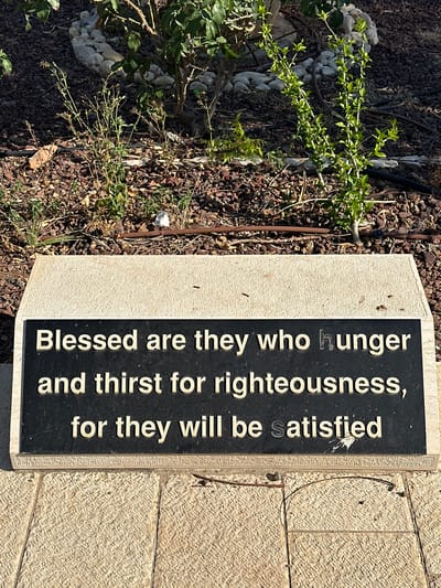 A black plaque with raised gold lettering is embedded in a beige stone base. The plaque reads: "Blessed are they who hunger and thirst for righteousness, for they will be satisfied." The plaque is situated outdoors on a cobbled walkway, with mulch, small plants, and rocks visible behind it, suggesting a garden or landscaped area. The overall setting implies a public space in Emek HaYarden Regional Council, Israel, possibly a park or memorial.