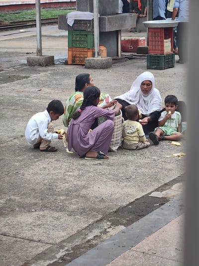 A group of people, including women and children, are sitting on a paved surface at what appears to be a train station. In the foreground, a young boy in a white shirt and tan pants kneels and holds a piece of banana. To his right, a young child wearing a light green outfit is eating a banana. A woman wearing a white headscarf and black garment is seated, interacting with the children. Another woman, wearing a patterned shirt and dark pants, is also seated. In the background, there are stacked crates, a Coca-Cola cooler, and train tracks, suggesting the location is a railway station. The overall environment appears to be an outdoor public space. The image shows an informal gathering of people, possibly a family, in a relatively unkempt urban setting.