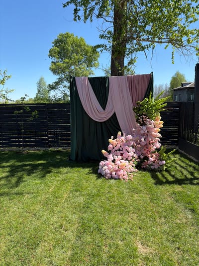 The image shows an outdoor decorative scene in Юг, Russia. A dark green and light pink draped backdrop is positioned against a dark wooden fence. The backdrop is adorned with arrangements of pink and green flowers. The scene is set on a lawn. A tree is on the left, and the sky is blue.