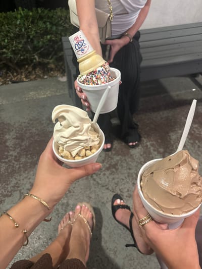 Three people are holding ice cream. One cup contains a chocolate ice cream and another has a vanilla ice cream and peanuts. The third cup has an ice cream cone with rainbow sprinkles. The location is likely Boca Raton, United States, based on the "Joy" ice cream cone which is located there.