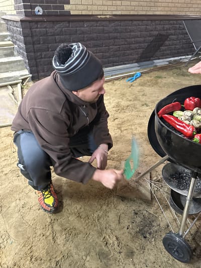 A man wearing a brown hooded jacket, black pants, and a gray and white beanie is crouched down and fanning a charcoal grill. The grill contains red bell peppers and small white vegetables, potentially mushrooms. The man is using a green fan to ignite the charcoal. He is wearing orange and black shoes. The ground appears to be dirt or sand. Part of a wall constructed of brick-like materials is visible in the background to the left. A pair of blue pliers are present on the ground behind the grill. The location appears to be outside, likely in слобода Старая, Russia.