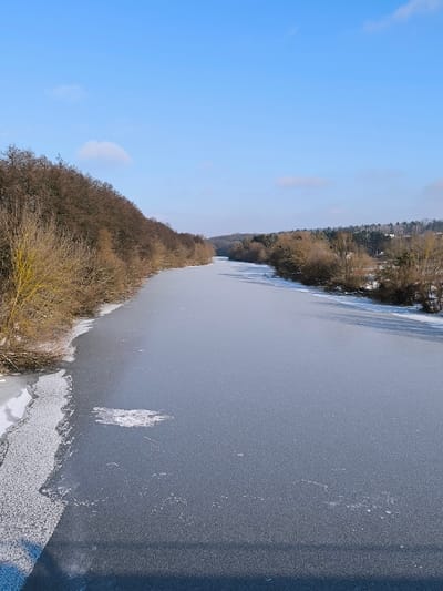 The scene depicts a frozen body of water in Prybuzke, Ukraine, viewed longitudinally under a clear blue sky with minimal scattered white clouds. The water's surface is completely covered by a solid, smooth, grayish-blue ice layer. An irregular patch of fragmented white ice or snow is visible on the frozen surface in the lower-left quadrant.
The left bank is densely covered with bare deciduous trees and shrubs, indicating a winter state, with snow accumulation visible along the water's edge and on the ground. The terrain on the left bank gradually ascends into a tree-covered slope. The right bank is also lined with bare deciduous trees and shrubs; behind them, in the mid-distance, several small buildings are partially visible amongst mixed coniferous and deciduous trees, suggesting a populated area. Snow is present along both banks.
A dark, angular shadow, originating from an unseen structure, extends across the lower portion of the frozen water surface in the foreground. No animate subjects or human activity are visible within the frame.