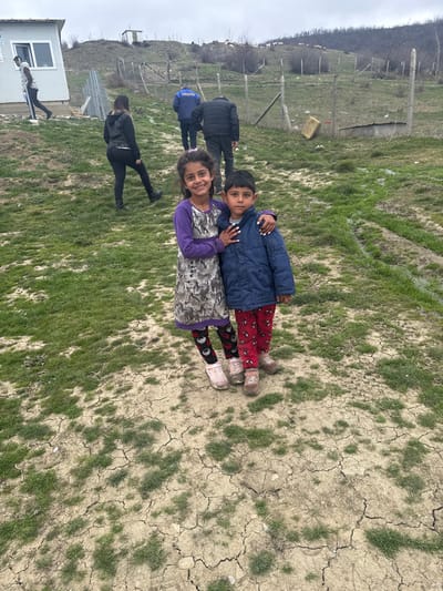 The image depicts an outdoor scene in Karageorgievo, Bulgaria, under an overcast sky. In the foreground, two children stand facing the camera. A girl, positioned on the left, wears a purple long-sleeved top under a grey and white patterned dress, black leggings with red motifs, and light pink shoes. She has long dark hair and her right arm is around the boy's shoulder. A boy, on the right, wears a dark blue padded jacket, red pants with white spider-like patterns, and light brown shoes; his left arm is around the girl's waist. The immediate ground is cracked, dry earth with scattered patches of green grass, transitioning to denser green growth further back.
Uphill from the children, a dirt path recedes into the background. A woman in a black jacket, black pants, and dark boots walks uphill on the left, followed by two men in the center-right. One man wears a blue jacket with "ГРАНИЧНА ПОЛИЦИЯ" (Border Police) visible on the back. To the far left, a male subject in a white vest is seen walking past a white modular building equipped with a single window. A wire fence on metal posts extends uphill through the mid-ground, separating the path from the vegetated hillside. A yellow plastic container hangs on the fence in the mid-right. The background features rolling hills with sparse, bare trees and shrubs, a small, rudimentary shack-like structure on a distant crest, and small white objects, consistent with sheep, on a higher slope.