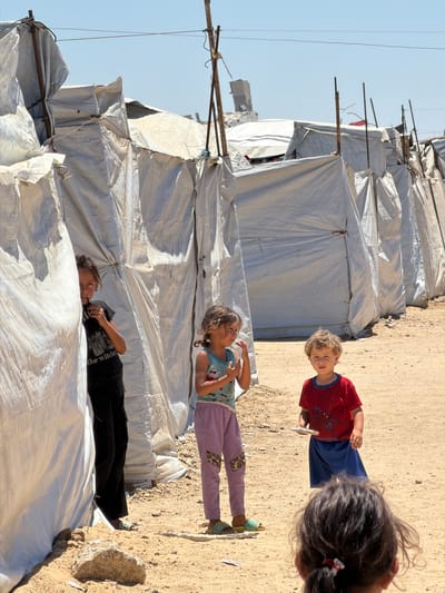 In the city of غزة, Unknown, a group of children are seen in a makeshift camp. The environment is characterized by temporary shelters made of white tarps stretched over metal poles. The ground is sandy and uneven, with scattered rocks and debris.

In the foreground, partially visible from behind, is a child with dark, curly hair. To the left, a girl is peeking out from behind a tarp, wearing a black t-shirt with a graphic print and black pants. She is holding something dark in her hand near her mouth. In the center, another girl with brown hair styled in a ponytail is wearing a light blue t-shirt with a print and purple patterned pants. She appears to be reaching for something or gesturing with her right hand. To the right, a young boy with blonde curly hair is wearing a red t-shirt with a Spider-Man print and blue shorts. He is holding a small white object, possibly a piece of food or a toy, in his hand. The children appear to be interacting or observing each other in the open area between the shelters. The sky is clear and blue, suggesting a bright, sunny day.