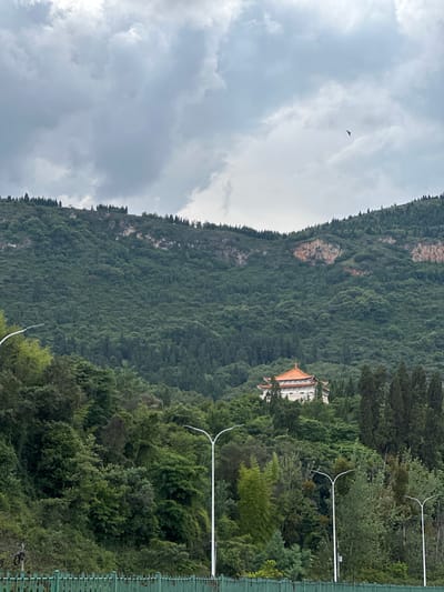 The image depicts a landscape scene in Xishan District, China, featuring a lush, tree-covered mountain slope under a cloudy sky. A traditional building with an orange-tiled roof is visible nestled amongst the trees on the mountainside, indicating a possible temple or historical structure. The foreground shows a green fence and several white lampposts, suggesting a road or public area. A solitary bird is captured in flight against the dramatic, overcast sky. The overall impression is one of natural beauty with a touch of architectural interest, characteristic of mountainous regions in China.