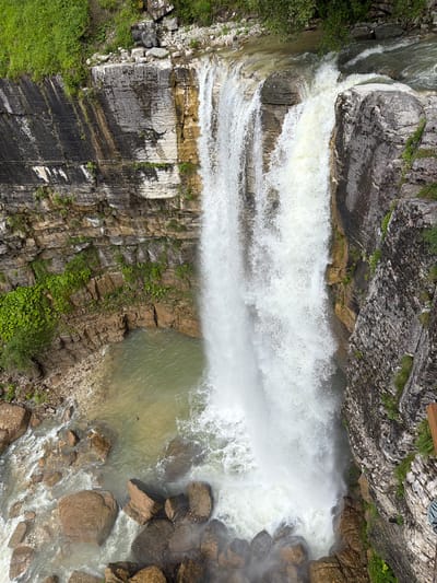 A high-angle, close-up, eye-level, wide shot captures a powerful, multi-tiered waterfall cascading down a rocky, layered cliff face. The water is a brilliant white, churning and frothing as it plunges into a pool of murky green water below. The surrounding rocks are a mix of dark gray and tan, with patches of vibrant green vegetation clinging to the cliff sides. The water at the top of the falls flows rapidly over a broad, uneven surface, creating smaller cascades before the main drop. The pool at the bottom is surrounded by large, brown boulders and smaller stones. The image is taken during the daytime, with ample natural light illuminating the scene. The location is the Kinchkhaperdi waterfall in Kinchkhaperdi, Georgia. There are no people or animals visible in the frame.