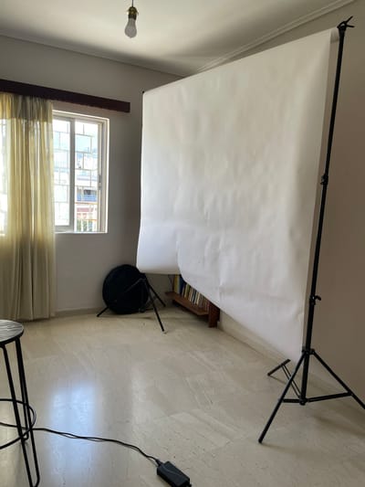 The image displays an indoor room, oriented as if rotated 90 degrees clockwise, showing a photography backdrop setup. When viewed upright (rotated 90 degrees counter-clockwise), the scene reveals a light-colored room with a white paper or fabric photography backdrop suspended from a black metal support system.
The backdrop system consists of a horizontal black bar held up by two vertical stands, one visible on the far left and the other on the far right. The white backdrop material hangs from this bar, cascading downwards and extending onto the floor, creating a seamless background. The backdrop appears somewhat wrinkled and has a few minor blemishes.
On the left side of the room, a window with multiple panes is visible towards the bottom. Light-colored, possibly sheer, curtains cover the lower half of the window, allowing daylight to filter through. Outside the window, glimpses of other buildings can be seen, indicating an urban or residential setting. Above and to the left of the window, a bare white light bulb in a simple, dark fixture hangs from the ceiling.
Towards the right side of the room, under the draped backdrop, a wooden bookshelf filled with books is partially visible. In front of the bookshelf, a small, dark object that resembles a collapsed tripod or a small, dark stool is present.
The floor of the room is covered with light-colored tiles, possibly marble or a similar material, exhibiting subtle patterns or veining. In the bottom right corner of the image, a black power adapter with a black cable extending from it is visible on the tiled floor. Part of a black metal leg, likely belonging to another stand or piece of photography equipment, is also seen in this area.
The walls are painted a light, neutral color. The overall impression is that of a functional space temporarily set up for photography, combining elements of a home environment with specialized equipment. The time of day appears to be daytime, judging by the light coming through the window.