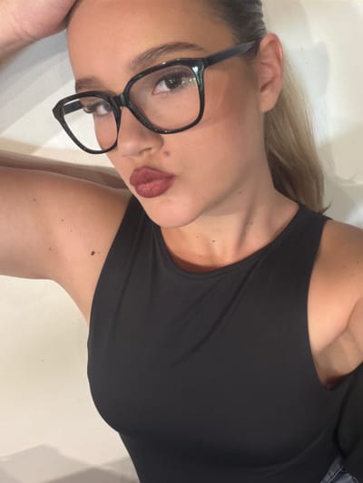 The media file shows a close-up, rotated shot of a woman lying down. She is wearing a black tank top and black-rimmed glasses. Her blonde hair is visible, and she has red lipstick on. The setting appears to be indoors, with a light-colored, textured surface underneath her. The lighting is soft and diffused, suggesting an indoor environment. There is no visible text in the image.