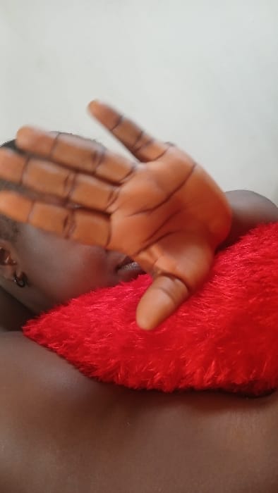 A close-up captures a person holding out a hand with distinct black lines drawn on the palm and fingers. Adjacent to the hand is a fluffy red object. The background is a mix of light gray and darker tones. The person's face is partially visible, showing an eye and parts of the lower face and neck, with an earring noticeable near the ear.
