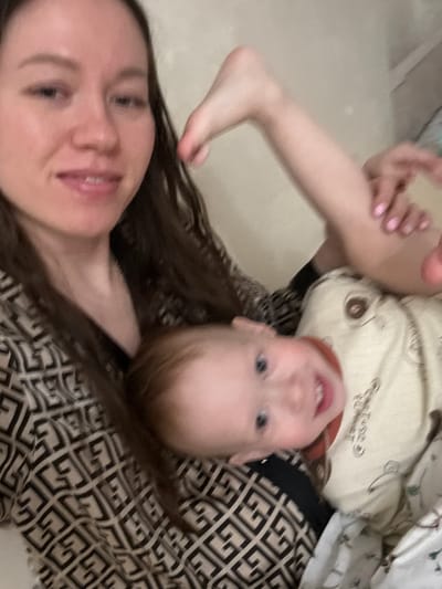 The image captures an indoor scene in the city of Shul'gino, Russia, featuring two individuals: an adult female and an infant. The adult female is positioned on the left side of the frame, presenting with fair skin, long dark brown hair, and a slight smile directed towards the camera. She wears a garment with a prominent repeating geometric pattern in contrasting light and dark shades. Her bare right foot and lower leg, with light pink painted toenails, are visible elevated in the upper right quadrant of the image, extending above the infant's head. The infant, located on the lower right, possesses light reddish-brown hair, blue eyes, and a wide smile revealing visible teeth. The infant is clad in a light-colored top featuring a dark, stylized pattern near its right shoulder. Both individuals are looking towards the camera. The background appears as an out-of-focus, plain, light-colored surface, suggesting an interior wall or soft furnishing. A section of white fabric with a faint, darker pattern, likely bedding, is visible in the lower right corner.