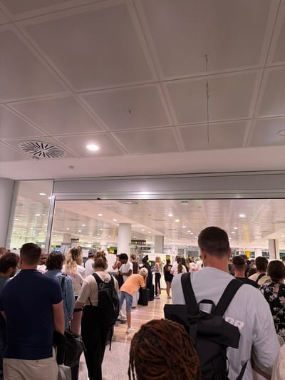 The image depicts a queue of people in what appears to be an airport terminal in Son Sant Joan, Spain. The environment is characterized by a white tiled ceiling with integrated lighting and ventilation systems. People are standing in line, facing away from the camera, suggesting they are waiting for a service such as check-in or security. They are dressed in casual attire. Some individuals are carrying luggage, including backpacks and suitcases. The overall scene conveys a sense of transit and anticipation.