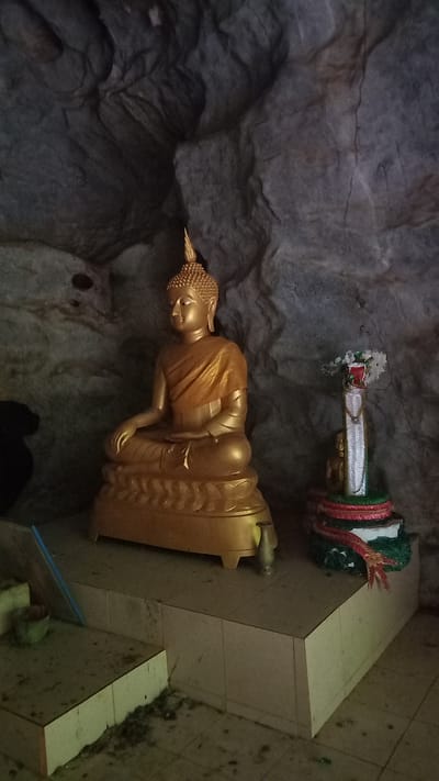 The photograph shows a golden Buddha statue inside a cave. The cave wall is rocky and dark. The Buddha sits on a decorated base, with offerings positioned behind it on a tiled platform. The image was taken at Ban Pak Ra, Thailand.