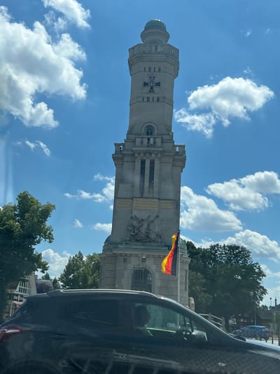 This image depicts a monument with a German flag flying in front of it. The monument is a tall, stone structure with inscriptions and decorative elements. The German flag, with its black, red, and yellow bands, is prominently displayed, suggesting a connection to German history or national pride. The scene appears to be set outdoors, with trees and a blue sky with white clouds visible in the background. A portion of a car is visible in the foreground, indicating the photo may have been taken from a vehicle.
Notable details include the years "1813" and "1915" inscribed on the monument, which likely refer to significant historical events. The monument itself is grand and imposing, suggesting it commemorates something of importance. The weather appears to be clear and sunny, with cumulus clouds scattered across the sky, indicating it's likely daytime. The location context provided is Großbeeren, Germany, which is significant as the Battle of Großbeeren took place in 1813, a key event in the Napoleonic Wars. This historical context, combined with the dates on the monument, strongly suggests the monument is related to this battle or a broader commemoration of German military history. No specific activities or events are directly depicted, other than the static display of the monument and flag. No visible emotions are conveyed in the image, as it focuses on the monument and its surroundings.