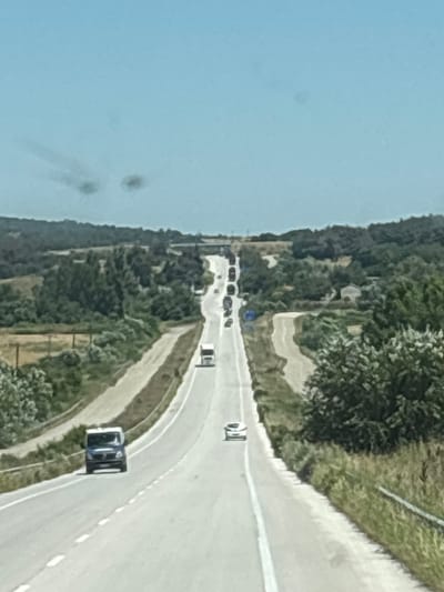 The image depicts a section of a highway in Λύρα, Greece, under a clear, pale blue sky. The road curves and ascends into the distance, flanked by lush green trees and vegetation on either side. Several vehicles are visible on the highway. In the foreground, a blue van is traveling away from the camera. Further up the road, a white car is also heading away. A white truck is seen further ahead, and a procession of other vehicles, including trucks and cars, stretches along the road in the distance. The landscape suggests a rural or semi-rural environment. The quality of the image is slightly blurred, possibly due to motion or lighting conditions.