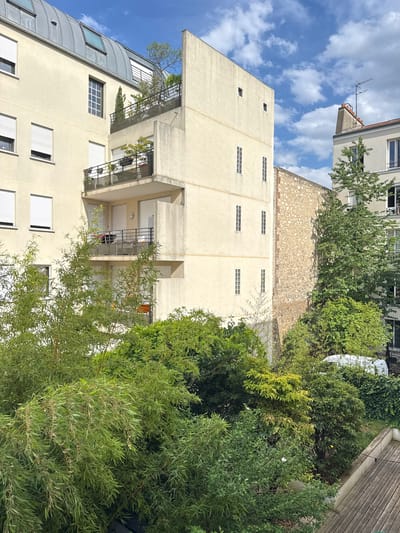 This is a photograph taken from a balcony, looking upwards and to the side. The scene is dominated by the architecture of apartment buildings and lush green foliage.
The primary subject is a cream-colored apartment building with multiple stories. It features a series of rectangular windows, some with white blinds, and balconies with black metal railings. One section of the building has a curved, grey metal exterior. Adjacent to this apartment building is a section of an older building with a textured stone facade and a tiled roof.
Lush green trees and bushes are abundant throughout the image, partially obscuring the buildings and creating a sense of a verdant urban environment. The trees vary in size and density, with some appearing to be bamboo or a similar slender-leafed variety.
The sky is visible in the upper left portion of the image and is a bright blue with scattered white clouds, suggesting it is daytime and the weather is fair. The sunlight casts shadows, indicating the time of day.
The location appears to be Montreuil, France, based on the context provided. There is no discernible activity or event taking place; it is a still scene capturing an urban landscape. No people are visible, and no text is present in the image. The overall impression is one of a bright, sunlit day in a densely vegetated urban area.