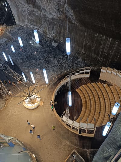 The image provides a high-angle view of the Turda Salt Mine in Turda, Romania. The vast underground salt cavern features a large, multi-tiered amphitheater with wooden seating and a stage area. The cavern walls are rough and striated, with natural salt formations visible. Several large, cylindrical fluorescent lights hang from the ceiling, illuminating the space.
In the foreground and mid-ground, people are scattered throughout the cavern floor, some walking, others gathered. A complex metal structure resembling a winch or a kinetic sculpture is situated on the sandy floor. A raised walkway made of wood and metal railings encircles a portion of the cavern, providing access to different areas. The overall impression is one of a unique, man-made structure integrated into a natural geological formation.
