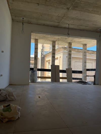 The image shows an interior view of an unfinished building under construction, likely an apartment or office space. The walls are bare concrete or plaster, and the ceiling is also exposed concrete, with electrical wires hanging down. The floor is tiled with large, pale tiles. A large opening in the wall looks out onto a street scene, with a building of classical architecture featuring columns and arched windows visible in the distance. There is a blue barrel and some construction materials, including large panels and stacks of what appear to be smaller tiles or planks, near the opening. A few bundles of fabric or sheeting are on the floor in the foreground. The scene is well-lit by natural light coming through the opening.