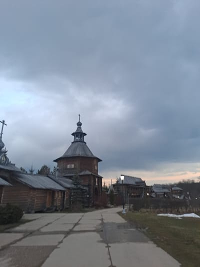 The image shows a view of the wooden architecture within the architectural and ethnographic museum in Suzdal, Russia, not Vzglyadnevo. The buildings are primarily constructed of wood, featuring pointed roofs and towers topped with Orthodox crosses. A concrete pathway is visible on the right side of the image. The sky is overcast with heavy, grey clouds. There are no visible people or actions depicted in the photograph. Patches of snow suggest late winter or early spring.