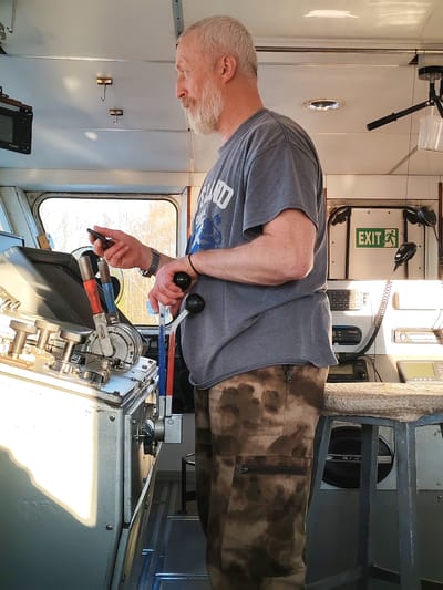 The image depicts an older man with gray hair and a beard standing inside what appears to be the control cabin of a train or boat. He is wearing a gray T-shirt and camouflage pants. He is holding a smartphone in his left hand and operating levers with his right hand. The controls are situated on a metallic console with various levers and gauges. Behind him, there is an "EXIT" sign and what appears to be radio communication equipment. There is a stool in the background. A window is visible, showing an exterior scene. The man's attention is focused on the instruments and the mobile phone. It is unknown whether the location is in Kurdiug, Russia.