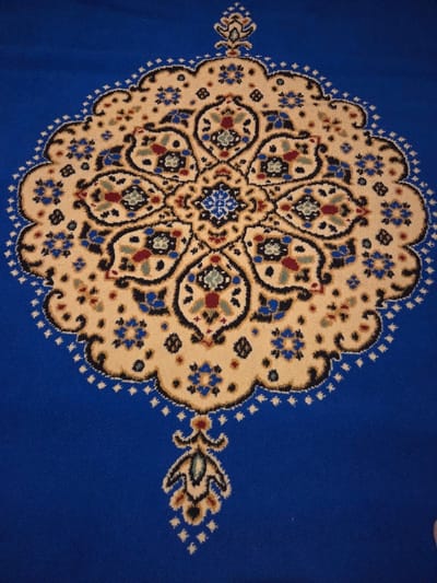 The image displays a close-up, top-down view of a patterned textile floor covering, specifically a carpet or rug. The dominant background color is a solid royal blue. Centrally featured is a large, intricate, symmetrical medallion design rendered primarily in beige/cream, outlined with a fine black border and small white/cream dots.
The medallion itself contains elaborate curvilinear and floral motifs. A smaller, star-like or multi-petal flower in shades of blue and black forms the absolute center. This is surrounded by radiating, lobed forms in beige, each containing detailed floral or leaf-like patterns in dark blue, light blue, red, green, black, and possibly gold/yellow. Smaller four-petal blue flowers are interspersed within the beige areas of the medallion.
Extending vertically from the top and bottom edges of the central medallion are smaller, elongated, stylized pendant designs, also featuring beige, black, and accent colors. The design appears consistent with traditional ornamental rug patterns. The carpet texture is uniform, suggesting a cut pile. No people, objects, or environmental elements beyond the carpet itself are visible. The specific location within Blainville-sur-Orne, France, is not visually identifiable in this image.