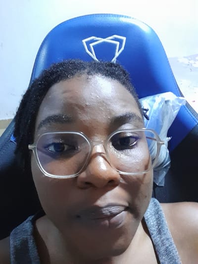 A person with dark hair and skin is wearing glasses and a gray top, sitting in a blue gaming chair. Plastic wrapping is visible near the headrest of the chair. The scene appears to be indoors, possibly in Umuigbekwo, Nigeria.