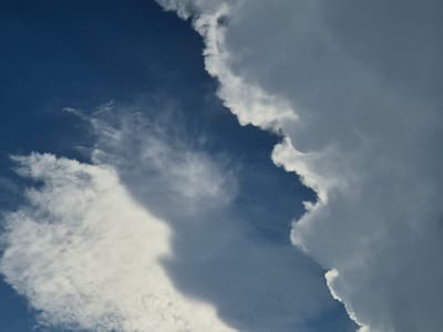 The image is a close-up, high-angle shot of the sky, featuring a dynamic interplay of dark blue sky and various cloud formations. On the right side of the frame, a large, dense, and overcast cloud dominates, casting a shadow and exhibiting a smooth, rounded edge where it meets the lighter sky. To the left, wispy, white cirrus clouds are scattered across the deep blue expanse, some appearing thin and feathery, while others are more substantial, suggesting a transition in atmospheric conditions. The lighting suggests it is daytime, with the sun likely positioned above or behind the main cloud mass. There are no people, structures, or identifiable landmarks present in the image. The image appears to have been taken from an elevated perspective, possibly from a building or a drone, looking upwards. No specific details about Rzeszów, Poland, can be discerned from this abstract depiction of the sky.