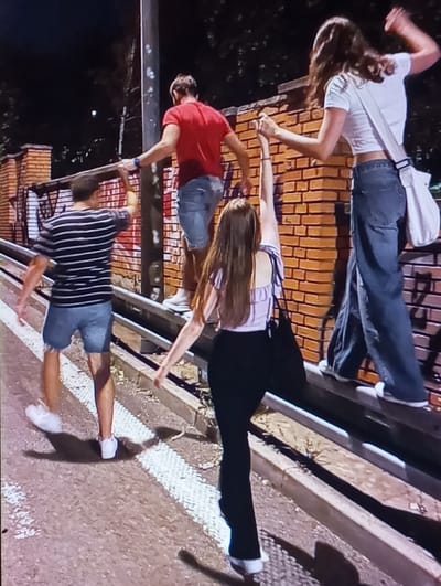 The image displays four individuals at night on a street in Baykaro, Uzbekistan. Two males and two females are present. A male, wearing a red t-shirt and blue denim shorts, stands on a low, silver-colored metal barrier. His left arm extends towards a dark vertical pole to his left. Adjacent to him on the same barrier is a female in a white t-shirt and blue wide-leg jeans. Her left hand is clasped with the raised right hand of a second female, who stands on the paved ground. This second female is dressed in a light-colored striped top, black flared pants, and carries a black shoulder bag. The fourth individual, a male in a horizontally striped t-shirt and blue denim shorts, stands on the paved ground to the left, with his left arm raised towards the vertical pole. Behind the metal barrier, a reddish-brown brick wall spans horizontally. The ground consists of dark asphalt or concrete, marked with a white painted line running parallel to the barrier. Dark foliage is visible above the brick wall against the night sky. The scene is illuminated by artificial light, casting distinct shadows.