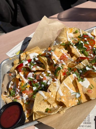 A high-angle shot reveals a tray of nachos, likely photographed outdoors in Huntington Beach, California. The nachos are topped with cheese sauce, tomatoes, onions, cilantro, and white cream sauce. A side of red salsa sits in a black plastic container. The tray is lined with brown paper. The photo is taken in daylight, with some shadows visible. Part of a person wearing a dark-colored jacket is visible in the lower left corner of the frame.