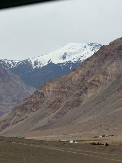 The image shows a vast, arid mountainous landscape under a cloudy sky. Dominating the background are towering, snow-capped mountains, their peaks shrouded in white. The slopes of the mountains are a mix of barren, rocky terrain with visible geological strata and patches of scree. In the mid-ground, a vast, flat valley floor stretches out, sparsely dotted with buildings. Two small, rectangular structures with green roofs are visible on the left side of the valley floor. Further to the right, a cluster of white buildings forms a small settlement, and beyond that, a few more scattered structures are discernible. The foreground consists of a dry, dirt track or road, with a rough, uneven surface. The overall impression is one of a remote, rugged, and sparsely populated mountainous region, characteristic of a high-altitude area like Zangla, India.
