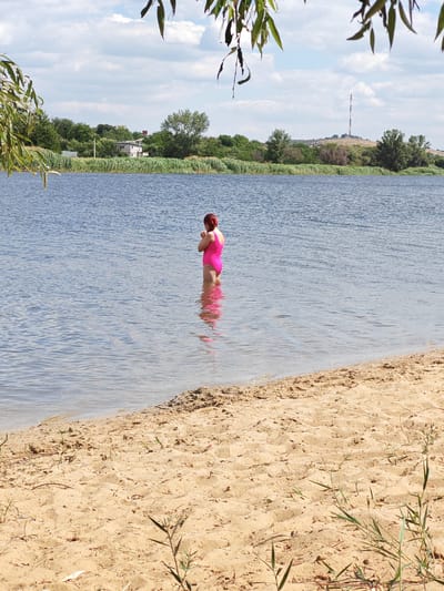 A person wearing a bright pink one-piece swimsuit stands in the shallow water of a wide river. The person's back is to the viewer, and they appear to be facing away from the camera, perhaps looking out at the water or performing an action with their hands near their face. The water is a deep blue and reflects the light, with small ripples visible. The riverbank in the foreground is a sandy beach with some sparse vegetation. In the background, across the river, is a lush green treeline and the embankment of the opposite shore. A few buildings are visible nestled within the trees. Further in the distance, a tall radio tower stands atop a hill. The sky is blue with scattered white clouds. The overall scene depicts a calm day at a riverside.
